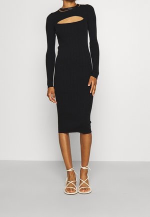 Osoba ubrana w czarną, długorękawową sukienkę midi z wycięciem na klatce piersiowej i białe sandały na wysokim obcasie z paskami, stojąca na szarej podłodze.