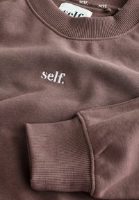 Bruine sweatshirt gemaakt van zachte stof, met een geribde ronde hals en manchetten, waarop het woord "self." in het wit geborduurd is.