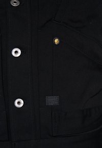 Černá tkanina s texturovanou vazbou, s použitím stříbrných a zlatých knoflíků a výraznou etiketou G-Star RAW. Funkční kapsy integrované do designu.