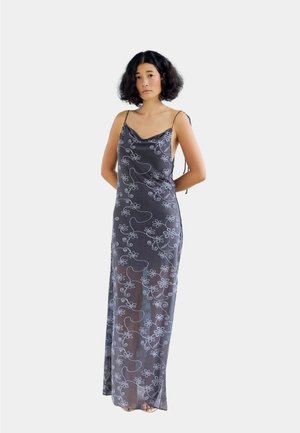 Femme aux cheveux bouclés foncés portant une robe noire transparente sans manches, longue jusqu'au sol, avec des motifs floraux blancs et fines bretelles.