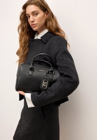 Črna usnjena torbica za čez ramo z zaobljeno obliko, dvema zgornjima ročajema, srebrnimi kovinskimi dodatki in odstranljivo etiketo. Model nosi temen plašč in črtasto srajco.