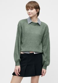 Joven con pelo corto castaño que lleva un suéter de punto verde sobre una camisa con cuello y una falda negra con plisados.