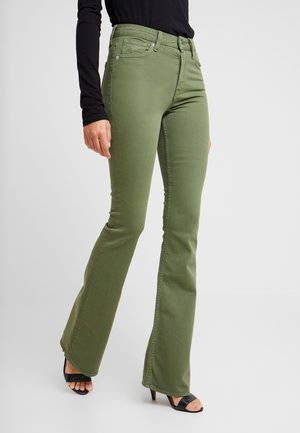 Kobieta ubrana w oliwkowe spodnie bootcut, czarną bluzkę z długim rękawem i czarne buty na obcasie z odkrytymi palcami stoi na jasnym tle.