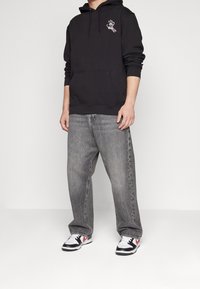 Černá mikina s kapucí a přední kapsou s vyšitým motivem ruky, v kombinaci se šedými džíny s širokými nohavicemi a černo-bílými teniskami.