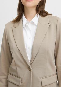 Beige blazer i strukturerad tyg med klandervärd krage, två framfickor och en enkel knappstängning, bärs över en vit skjorta.