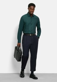 Camicia a maniche lunghe verde scuro, abbottonata, abbinata a pantaloni scuri, cintura nera e scarpe nere. Tiene in mano una valigetta in pelle nera.