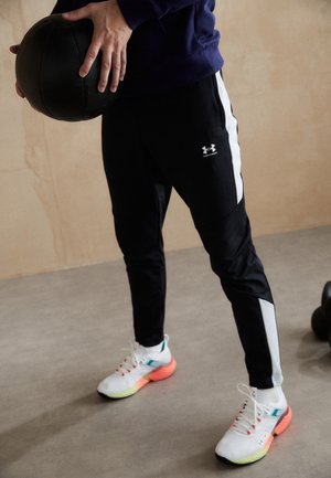 Personne en pantalon de survêtement noir et baskets blanches tenant un ballon médicinal noir, debout sur un sol gris devant un mur beige.