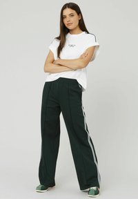T-shirt blanc à manches courtes avec du texte, associé à un pantalon large vert foncé avec des rayures blanches sur les côtés et une taille élastique. Chaussures vertes.