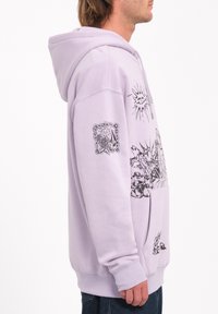 Haut à capuche violet clair avec une poche avant, comportant des motifs et des designs graphiques noirs sur la poitrine et la manche gauche. Tissu doux.