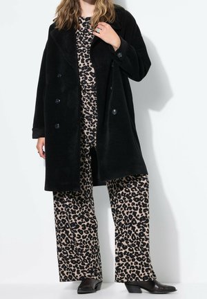 Femme portant un manteau noir à double boutonnage sur un pantalon large imprimé léopard beige et noir et un haut assorti avec des chaussures à talons noires.