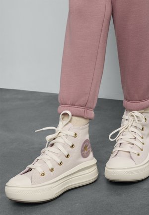 Sneakers typu high-top w jasnoróżowym płótnie z złotymi oczkami, z białą gumową podeszwą i okrągłym fioletowym logo po boku.