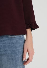 Eine Hand ruht neben blauen Jeans und trägt ein dunkelburgunderfarbenes Langarmshirt mit rüschierten Manschetten.