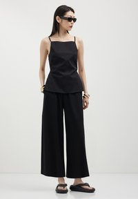 Donna che indossa un top senza maniche nero e pantaloni larghi, occhiali da sole neri, bracciali dorati e sandali neri con zeppa, in piedi su uno sfondo bianco.