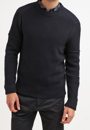 Homme portant un pull en maille texturé bleu marine foncé avec des manches à motifs et un col rond festonné, par-dessus une chemise à fleurs.