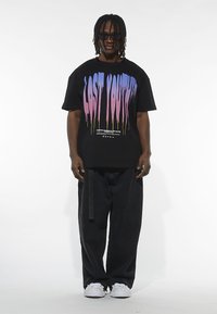 T-shirt nera oversize con testo gocciolante in gradiente viola e rosa. Abbinata a pantaloni larghi neri e sneakers bianche.