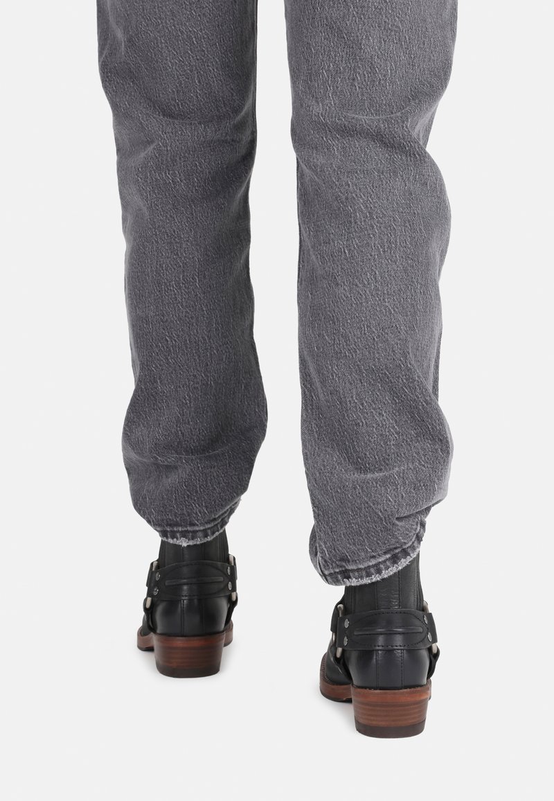 Schwarze Lederstiefeletten mit einem Holzabsatz und metallischen Akzenten, kombiniert mit grauen Jeans, die eine strukturierte, ausgebleichte Optik aufweisen.