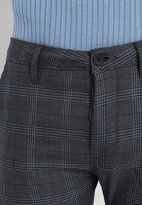 Šedé kalhoty se vzorem, které mají kostkovaný design s modrými akcenty, zapínání na knoflík a strukturovanou látku poblíž pasu.