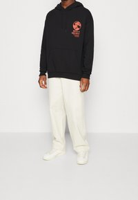Sudadera negra con un bolsillo frontal y cordones, con gráficos y texto en rojo. Combinada con pantalones relajados de un tono beige claro y zapatillas blancas.
