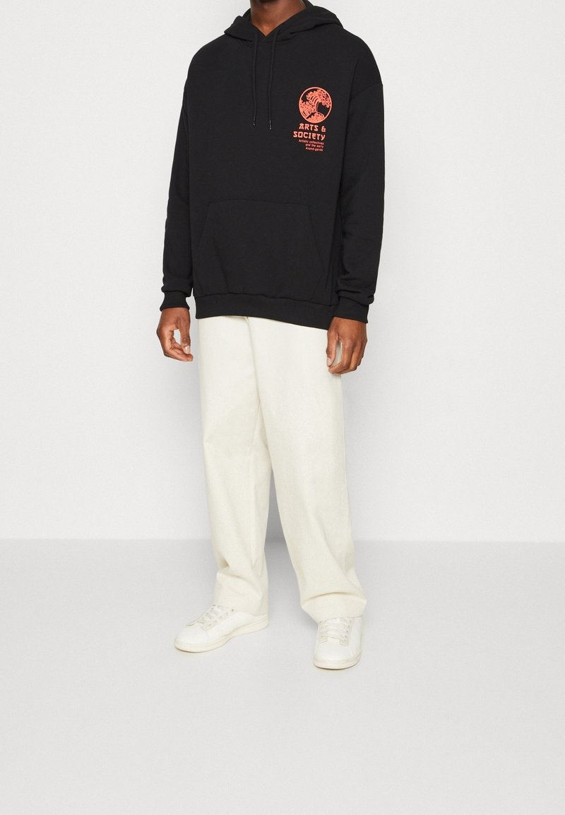 Sudadera negra con un bolsillo frontal y cordones, con gráficos y texto en rojo. Combinada con pantalones relajados de un tono beige claro y zapatillas blancas.