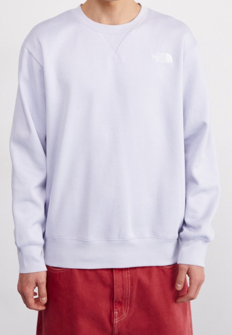 Personne portant un sweat-shirt violet clair avec un petit logo blanc sur la poitrine gauche et un pantalon rouge, debout devant un fond uni.