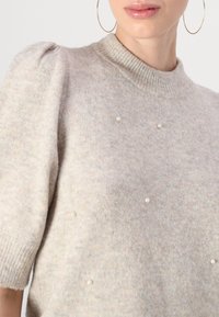 Sweater cinza tricotado com mangas curtas bufantes, decote canelado e pérolas falsas decorativas espalhadas pela frente. Textura macia.
