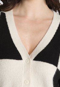 V-Ausschnitt-Strickjacke mit einem schwarz- und cremefarbenen geometrischen Muster, gerippter Textur und Knopfverschluss vorne.