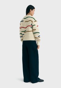 Flauschiger cremefarbener Pullover mit mehrfarbigen horizontalen Streifen, strukturiertem Strickmuster und geripptem Saum. Kombiniert mit dunklen, weit geschnittenen Hosen.