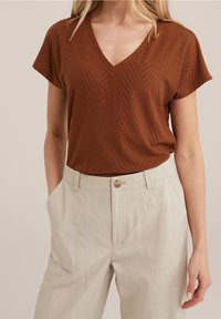 Brun v-ringad topp med korta ärmar och ett chevronmönster, i kombination med ljusbeiga linnebyxor med sidofickor och knappstängning.