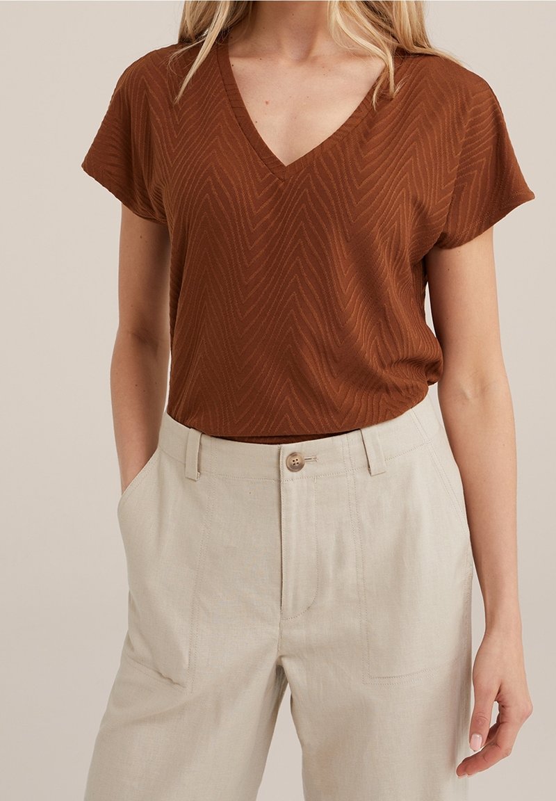 Brun v-ringad topp med korta ärmar och ett chevronmönster, i kombination med ljusbeiga linnebyxor med sidofickor och knappstängning.