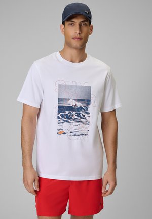 Mężczyzna w białej koszulce z grafiką oceanu i napisem "SUMMER SEASON", czerwonych szortach i granatowej czapce, stoi na szarym tle.