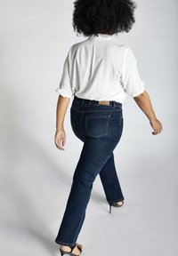Jeans de denim azul oscuro con un ajuste slim, combinados con una blusa blanca y sandalias de tacón negro, exhibiendo una silueta curvada.