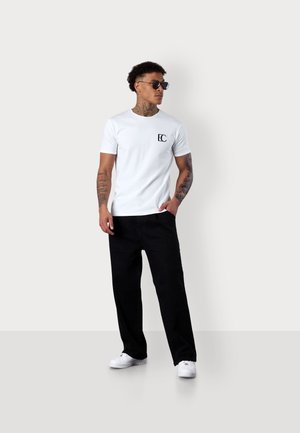 White cotton t-shirt with black logo on chest, paired with black wide-leg pants and white sneakers, model wearing sunglasses.