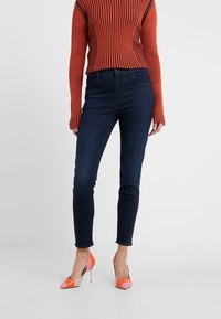 Vaqueros ajustados de color azul oscuro hechos de mezclilla, combinados con un suéter de manga larga acanalado en rojo y marrón y zapatos de tacón de colores múltiples.