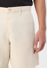 Beige shorts valmistettu teksuuripinnasta, jossa on yksi etutasku, nappikiinnityksellä varustettu vyötärö ja sivusaumayksityiskohtia.