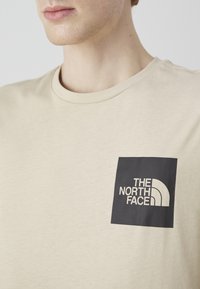 Camiseta de cuello redondo color beige con mangas cortas, que presenta un parche cuadrado negro con el logo de "THE NORTH FACE" en blanco. Textura de tela suave.
