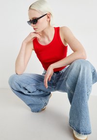 Röd randig linne med vida, ljusblå denimjeans, texturerad tyg och svarta solglasögon. Modellen sitter i knä med handen på hakan.