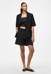 Blazer preto sob medida com mangas curtas sobre um top cropped preto e calções correspondentes. Tecido suave com um corte estruturado e detalhes mínimos.
