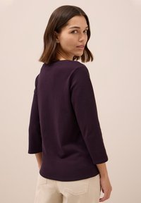 Maglia lunga manica color viola scuro con scollatura rotonda. Tessuto liscio e design aderente, vista da dietro, abbinata a pantaloni color beige chiaro.