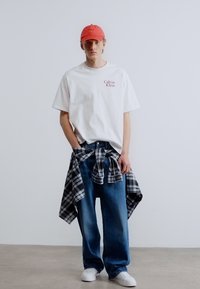 Άσπρη μπλούζα Calvin Klein, χαλαρό τζιν τζιν με μπλε χρώμα, ασπρόμαυρο καρό πουκάμισο δεμένο στη μέση, κόκκινο καπέλο, λευκά αθλητικά παπούτσια.