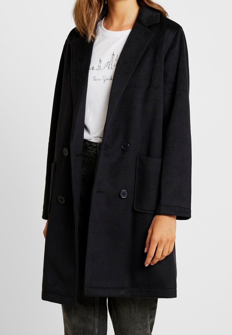 Femme portant un manteau noir croisé sur un t-shirt blanc avec un croquis de la silhouette de New York et un pantalon noir.