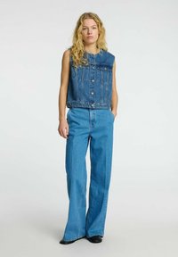 Chaleco de denim sin mangas, con dos bolsillos en el pecho, cierre de botones, combinado con pantalones de denim de pierna ancha de cintura alta en un tono azul claro.