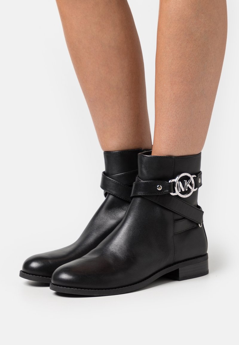 Bottines en cuir noir avec des talons bas, des sangles enroulées et une boucle ronde en argent avec le logo MK, portées sur des jambes nues.