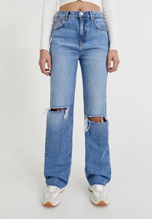 Person trägt hoch taillierte, hellblaue Jeans mit Rissen an beiden Knien, ein weißes, kurzes Langarmshirt und weiße Turnschuhe.