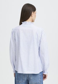 Camicia bianca con righe verticali blu, collo arricciato e polsini raccolti. Il tessuto è leggero con una texture liscia.