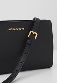 Svart crossbodyväska i syntetiskt läder med en guldig "MICHAEL KORS"-logotyp, justerbar rem och slät textur.
