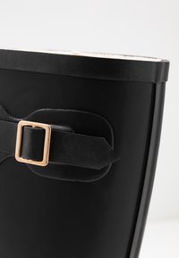 Botte en caoutchouc noire avec un détail en boucle sur le côté et une surface texturée. L'intérieur est doublé, avec un bord supérieur lisse.