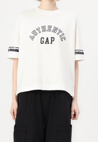 Bílý bavlněný tričko s potiskem "AUTENTICKÝ GAP" v černé barvě, krátké rukávy s tmavými pruhy na každém. Volný střih.