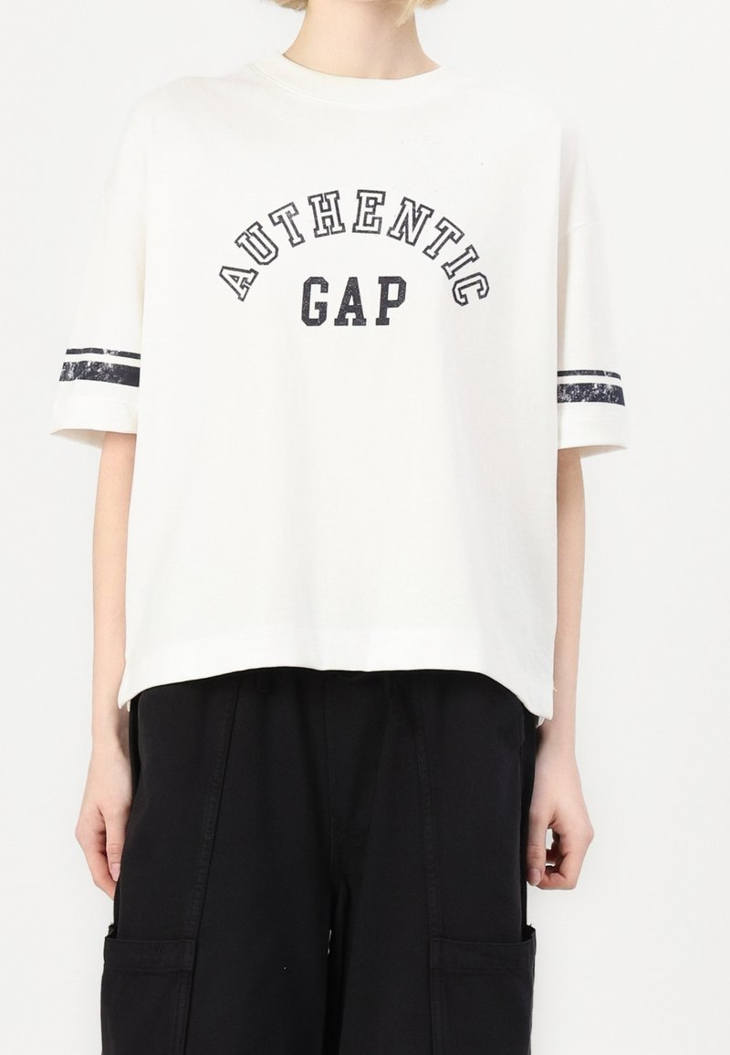 Bílý bavlněný tričko s potiskem "AUTENTICKÝ GAP" v černé barvě, krátké rukávy s tmavými pruhy na každém. Volný střih.