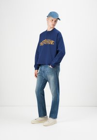 Marineblå hettegenser med gul tekst, lyseblå caps, lyseblå jeans og beige sneakers. Uformell antrekk med avslappet passform og minimalistisk design.