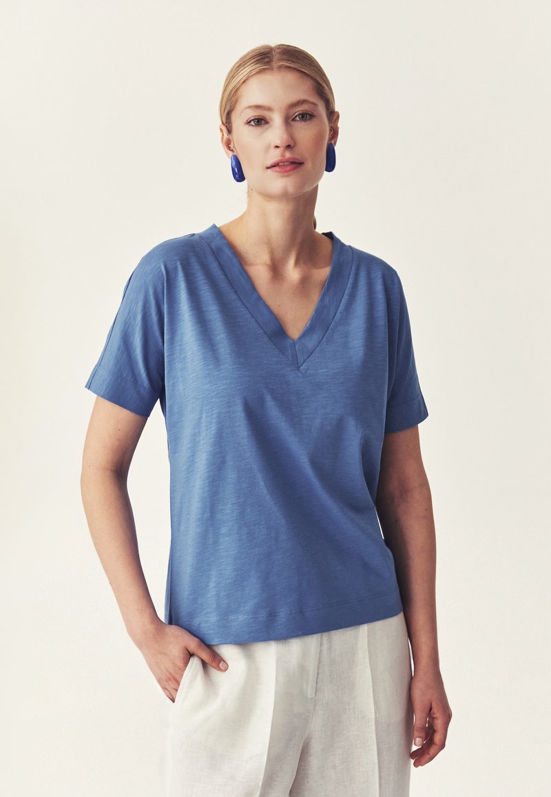 Femme aux cheveux blonds portant un t-shirt bleu à col en V et manches courtes, un pantalon blanc et des boucles d'oreilles ovales bleues, debout avec une main dans la poche.